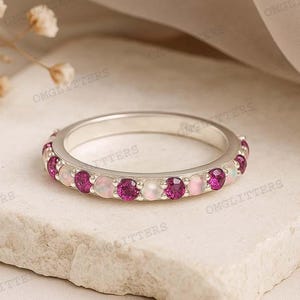 May include: A silver ring with alternating pink and white gemstones. The ring is set on a light-colored stone surface. The gemstones are round and evenly spaced around the band. The ring is a piece of jewelry.