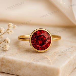 Könnte beinhalten: Ein goldener Ring mit einem großen, runden, facettierten roten Edelstein. Der Ring liegt auf einer Marmoroberfläche mit einem weichen, neutralen Hintergrund. Das Wort "OMGLITTERS" ist im Bild zu sehen.