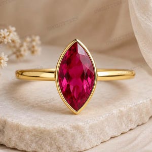 May include: A gold ring featuring a marquise-cut vibrant red gemstone set in a simple polished band. The faceted stone is positioned vertically, showcasing deep crimson tones and a clear, reflective surface held by a thin, smooth gold metal setting.