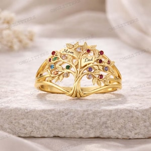 Puede incluir: Un anillo dorado con un diseño de árbol de la vida. El árbol tiene hojas de piedras preciosas de colores y las palabras "Mom" y "Dad" grabadas en el tronco. El anillo está sobre una superficie clara.