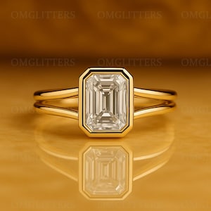 Puede incluir: Un anillo de oro con una gran piedra preciosa clara de forma rectangular. La piedra está engastada en un bisel de oro y tiene un diseño de talla escalonada. El anillo tiene un diseño de caña dividida, con dos bandas que se fusionan en la parte posterior.