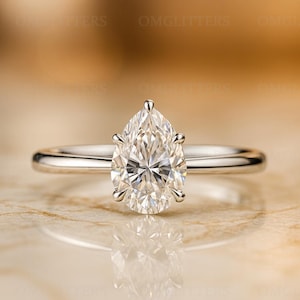 May include: A solitaire engagement ring featuring a pear-shaped diamond set on a thin, polished silver band. The diamond is clear and faceted, reflecting light. The ring is displayed on a reflective surface.