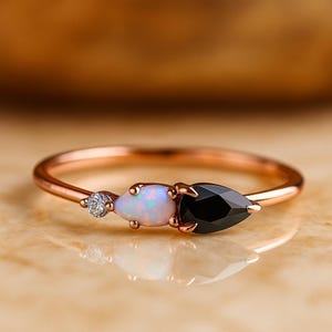 Può includere: Anello in oro rosa con un piccolo diamante, un'opale a forma di pera e un onice nero a forma di goccia. L'anello ha una fascia sottile e delicata, con le pietre preziose vicine. Le pietre preziose sono il punto focale dell'anello.
