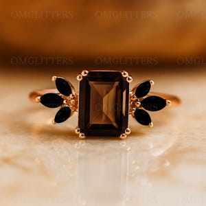 May include: A rose gold ring featuring a rectangular, brown gemstone center. Flanking the center stone are clusters of black, leaf-shaped stones. The ring has a delicate, elegant design, suitable for special occasions.