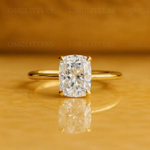 Puede incluir: Un anillo de oro con una gran gema transparente de talla cuadrada. La gema está engastada con cuatro garras y refleja la luz. La banda del anillo es fina y pulida, con un diseño elegante y sencillo.