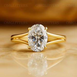 Könnte beinhalten: Ein goldener Ring mit einem großen, oval geschliffenen Diamanten. Der Diamant ist der Mittelpunkt und reflektiert das Licht. Der Ring hat ein geteiltes Schaftdesign, was seine Eleganz unterstreicht. Der Ring liegt auf einer reflektierenden Oberfläche.