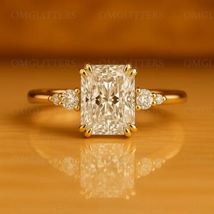 Può includere: Anello di fidanzamento in oro con un diamante rettangolare al centro. L'anello presenta diamanti rotondi più piccoli su entrambi i lati della pietra principale. L'anello è esposto su una superficie riflettente.