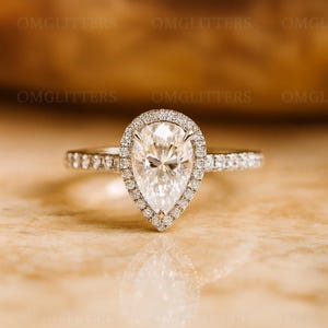 以下が含まれることがあります： 洋梨型のダイヤモンドをあしらった指輪。中央の石の周りには小さなダイヤモンドのハローが施されています。バンドにも小さなダイヤモンドがちりばめられています。反射する表面に置かれています。