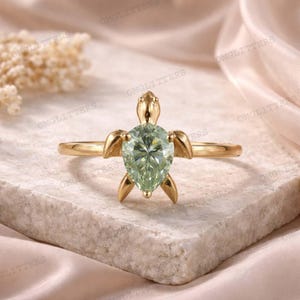 May include: A gold-colored ring featuring a turtle design. The turtle's body is a pear-shaped, light green gemstone. The ring's band and turtle details are gold-toned, creating a nature-inspired jewelry piece.