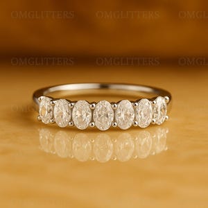 Op de afbeelding: Een zilveren ring met zeven ovale edelstenen in een rij. De heldere edelstenen schitteren en reflecteren het licht. De ring wordt getoond op een reflecterend oppervlak.