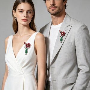 May include: A woman in a white dress and a man in a gray suit each wear a floral brooch. The brooches feature a red flower, white accents, and a green base. The woman's dress has a V-neck.