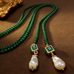 Op de afbeelding: Een groene kralenketting met twee strengen, met vierkante groene edelsteenaccenten in gouden zettingen. Elke streng eindigt met een grote, druppelvormige parelhanger met gouden en kristallen details. De ketting wordt gepresenteerd op een bruin oppervlak.