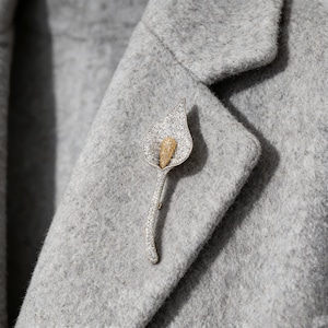 May include: A silver-toned calla lily brooch with a gold-colored center, encrusted with small, sparkling stones. The brooch is pinned to a gray wool coat, showcasing its elegant design and craftsmanship.