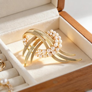 May include: A gold brooch with a layered design, featuring a central cluster of small pearls. The brooch has a textured, feather-like pattern and is displayed in a wooden jewellery box with a soft, white interior.