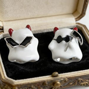 May include: A pair of white ghost-shaped earrings with red horns and silver detailing. One features black bat-wing elements, the other wears sunglasses and holds a small scythe. Displayed in a vintage jewellery box.