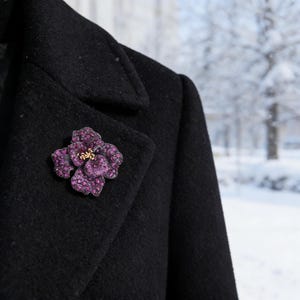 May include: A dark purple flower brooch with a gold centre, pinned on a black coat. The brooch is made of small, sparkling stones. The background shows a snowy, winter scene.
