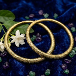 May include: Two gold bangles with an etched, decorative pattern. The bangles are set against a dark blue backdrop, with white flowers, purple and green gemstones. The image is a close-up of the jewellery.