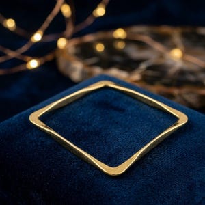 Puede incluir: Una pulsera cuadrada de color dorado con lados suavemente curvados. La pulsera se muestra sobre una superficie de terciopelo azul oscuro, con luces de cadena borrosas en el fondo.