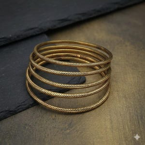 May include: A stack of six gold-colored bangles. Each bangle has a series of small, evenly spaced perforations. The bangles are arranged in a spiral, with the top bangle slightly offset. The background is a dark gray and brown surface.