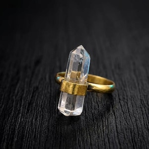 May include: A gold ring with a clear quartz crystal. The crystal is held vertically by a gold band. The ring's band is also gold. The background is a dark, textured surface.