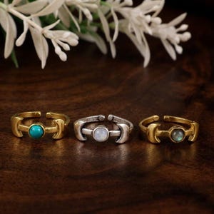 May include: Three adjustable rings featuring a crescent moon design on each side of a central round gemstone. The left ring is gold-toned with a turquoise stone, the center is silver-toned with a white stone, and the right is gold-toned with a grey stone.