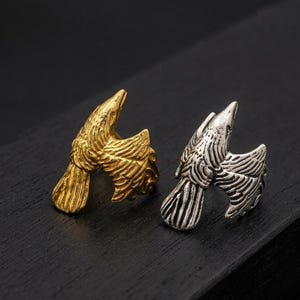 May include: Two rings, one gold-colored and one silver-colored, each featuring a stylized eagle design. The eagles are depicted with outstretched wings and detailed feathering, set against a dark background.