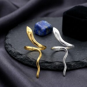 Puede incluir: Dos anillos con forma de serpiente, uno dorado y otro plateado, se muestran sobre una superficie oscura y texturizada. Los anillos presentan un diseño en espiral con una cabeza de serpiente en la parte superior. Una piedra azul y un joyero negro están en el fondo.
