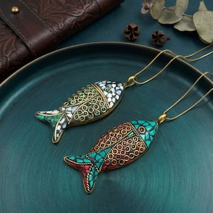 May include: Two fish-shaped pendant necklaces. The pendants are adorned with colorful mosaic-style inlays of turquoise, red, green, and white. The fish have gold-colored metal outlines and are suspended from delicate gold chains. The background is a teal-colored tray.