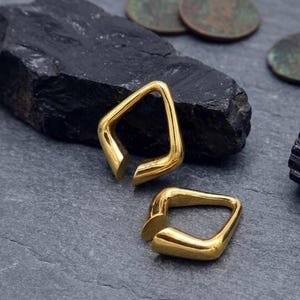 May include: Two gold-toned ear weights with a smooth, polished finish and a distinct geometric, open-ended square shape. These metallic pieces feature rounded edges and a continuous, solid construction, presenting a reflective surface.