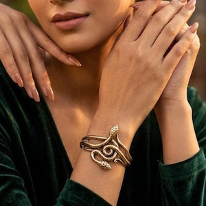 May include: A gold-toned metal cuff bracelet shaped like a coiled snake. The bracelet features a textured, scale-like surface pattern that wraps around the wrist, with the snake's head and tail meeting at the front in a detailed, overlapping design.