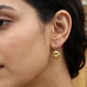 Puede incluir: Un pendiente de tono dorado que presenta un pequeño dije de concha marina texturizada suspendido de un gancho simple. La concha tiene un diseño de superficie acanalada y un acabado metálico pulido, colgando directamente del lóbulo.