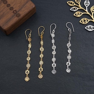Può includere: Quattro paia di orecchini pendenti con simboli dei chakra in tonalità oro e argento sono esposti su una superficie nera. Gli orecchini sono lunghi e delicati, con una serie di forme circolari e geometriche. Sullo sfondo, una scatola di legno con simboli dei chakra incisi.
