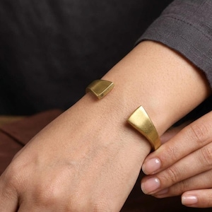 Pode incluir: Uma pulseira de punho dourada com um design geométrico. A pulseira aberta apresenta duas extremidades retangulares ligeiramente anguladas. A pulseira é usada no pulso, mostrando seu estilo simples, mas elegante.