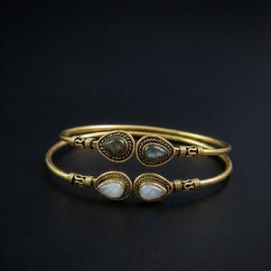 May include: Two open-ended gold-toned metal cuff bracelets featuring teardrop-shaped gemstones at each terminal. The top bracelet displays dark, iridescent stones, while the bottom bracelet features smooth, translucent white stones.