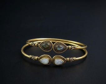 May include: Two open-ended gold-toned metal cuff bracelets featuring teardrop-shaped gemstones at each terminal. The top bracelet displays dark, iridescent stones, while the bottom bracelet features smooth, translucent white stones.