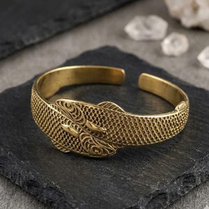 Puede incluir: Un brazalete dorado con diseño de pez. El brazalete presenta dos cabezas de pez en el centro, con patrones detallados de escamas y aletas. El diseño abierto facilita su uso.