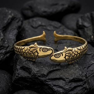May include: A gold-colored cuff bracelet featuring two fish heads facing each other. The fish have detailed scales and eye designs. The bracelet is open-ended and rests on a dark, textured surface.
