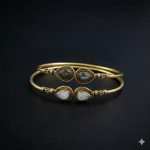 May include: Two gold-colored bangles with teardrop-shaped gemstones. One bangle pair features green and blue stones, the other pair has white stones. The bracelets have intricate detailing and are set against a black background.