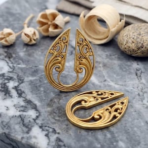 May include: A pair of gold-toned ear weights featuring an open teardrop shape with intricate scrollwork patterns. One piece stands upright while the other lies flat, both showcasing detailed metal filigree designs with a polished finish.