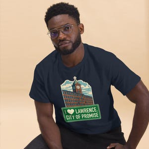 May include: Navy blue t-shirt featuring a graphic of a brick building with a clock tower and the text "I ❤️ Lawrence, City of Promise" on a green sign. The shirt is a short-sleeved crew neck.