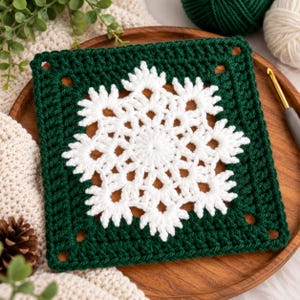 May include: A square crochet granny square featuring a white snowflake motif centered on a dark forest green background. The intricate white yarn pattern creates a star-like design with pointed edges, framed by a solid dark green border.
