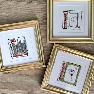 Może przedstawiać: Trzy kwadratowe, złote ramki z białym passe-partout i ilustracjami. Jedna przedstawia panoramę NYC, druga pudełko zapałek z napisem Lucky You, a trzecia kieliszek do martini z napisem It's Tini Time.