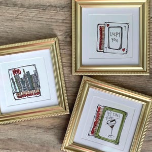 May include: Three square gold-toned frames containing white matting and illustrations. One features a NYC skyline, another a matchbook with the text Lucky You, and the third a matchbook with a martini glass and the text It's Tini Time.