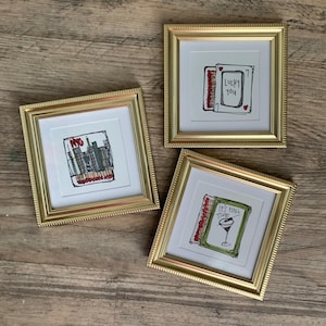 May include: Three framed art prints with gold frames. One print features a NYC skyline, another says "Lucky You", and the third says "It's Fine Time". The prints have a white matting and are on a wooden surface.