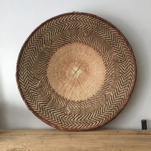 May include: A large, round woven basket with a natural, earthy aesthetic. The basket features a central light tan circle, surrounded by a brown and tan chevron pattern. The rim is a darker brown, creating a defined edge. The basket is likely handmade.