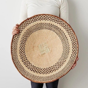 May include: A large, round woven basket with a natural beige center and a geometric brown and beige pattern around the edges. The basket has a dark brown rim and is held by a person.
