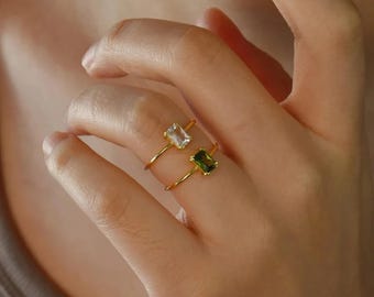 May include: Two gold rings, each with a rectangular gemstone. One ring features a clear stone, the other a green stone. The rings are displayed on a hand, showcasing the jewelry's design and color.
