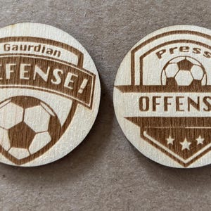 Pode incluir: Duas fichas de madeira com tema de futebol. Uma diz "Gaurdian DEFENSE!" com um gráfico de bola de futebol. A outra diz "Press OFFENSE!" com um gráfico de bola de futebol e três estrelas. As fichas são castanhas claras.