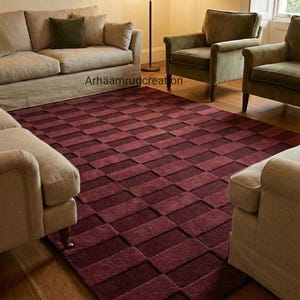 May include: A rectangular, burgundy area rug with a geometric pattern of alternating shades of red. The rug is placed in a living room with beige sofas and green armchairs. The text "Arhaamrugcreation" is visible.
