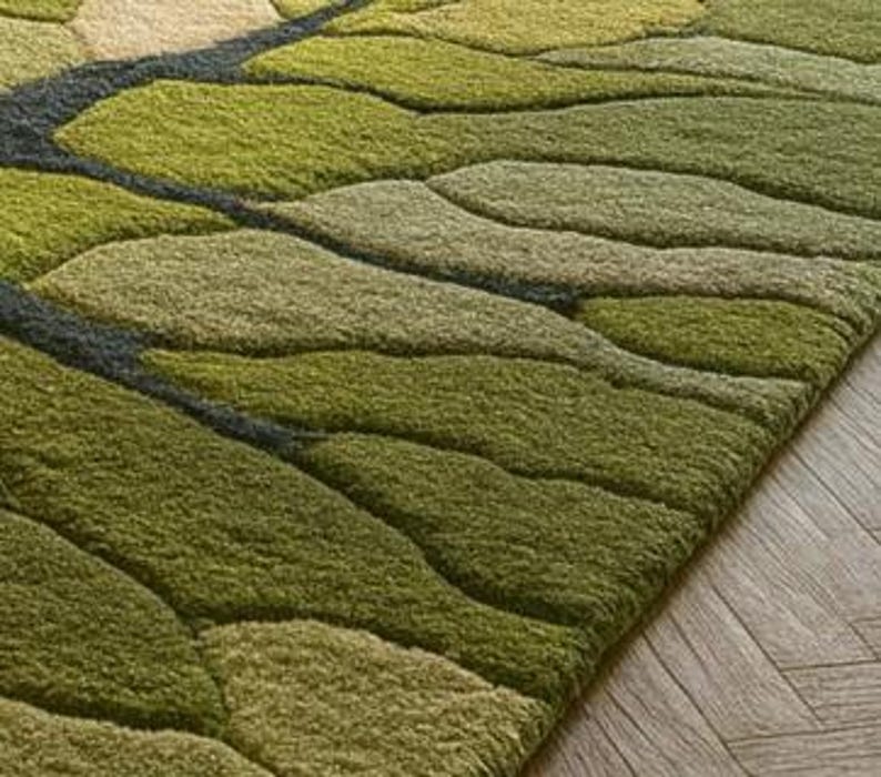 May include: A green area rug with a leaf-like pattern. The rug features varying shades of green, from light to dark, with a dark gray border. The rug is placed on a wooden floor.